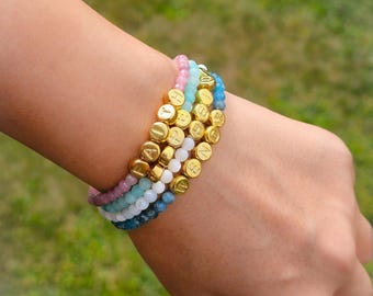 May include: A stack of beaded bracelets in shades of pink, blue, white, and gold. The gold beads spell out the word "HOPE". The bracelets are worn on a wrist against a blurred green background.