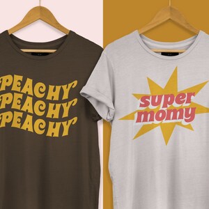 以下が含まれることがあります： レトロなグラフィックがプリントされたTシャツ2枚。1枚は茶色のTシャツで、黄色で「Peachy Peachy Peachy」と書いてある。もう1枚は白いTシャツで、黄色の星形の中に赤と黄色の「super mommy」と書いてある。