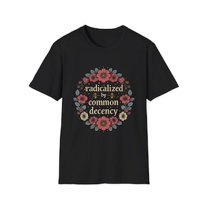 May include: Black t-shirt featuring a floral wreath design with the words "radicalized by common decency" in a stylized font. The wreath includes pink, red, and yellow flowers with green leaves.