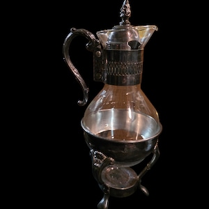 Puede incluir: Una cafetera vintage plateada con una jarra de cristal transparente. La cafetera presenta un asa ornamentada, una banda decorativa alrededor del centro y un pequeño soporte con tres patas. La tapa está rematada con un adorno decorativo.