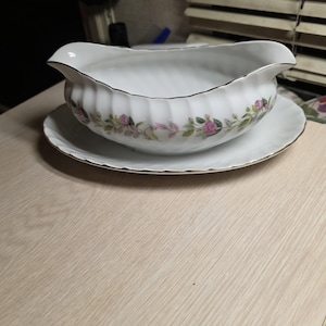 May include: White porcelain gravy boat and matching saucer with gold trim. The boat has a scalloped edge and a floral design with pink roses and green leaves. The set is on a light-colored wooden surface.