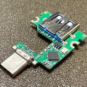 Puede incluir: Primer plano de una placa de circuito verde con un puerto USB y un puerto USB-C. La placa tiene varios componentes electrónicos, incluidos chips y conectores. El puerto USB es plateado y azul, y el puerto USB-C es plateado.