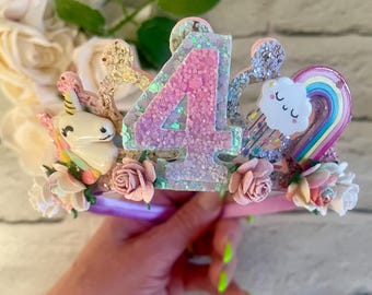 Könnte beinhalten: Ein rosa-lila glitzernder Haarreifen mit einer großen pinkfarbenen Zahl vier, einem Einhorn, einem Regenbogen und einer Wolke mit einem Smiley. Der Haarreifen ist mit kleinen rosa Blüten verziert.