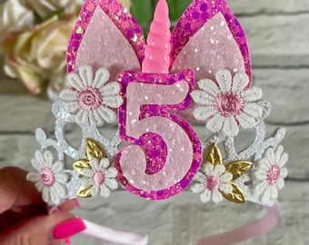 May include: White headband with a pink unicorn horn and ears, decorated with white lace flowers and gold leaves. The number 5 is in the center, with pink glitter and a white background.