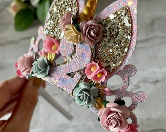 May include: A purple and gold glitter unicorn headband with a white headband and pink, white, and green flowers.
