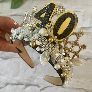 May include: A black headband adorned with a gold and glittery "40" numeral, pearls, and decorative leaves. The crown-like design features gold and silver accents, creating a festive accessory for a milestone birthday celebration.