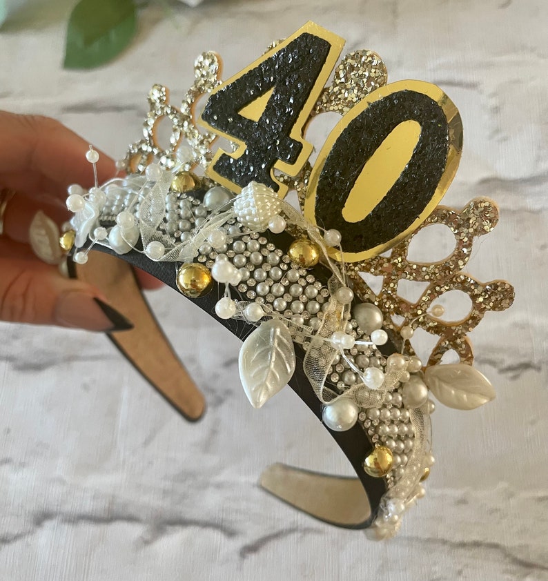 May include: A black headband adorned with a gold and glittery "40" numeral, pearls, and decorative leaves. The crown-like design features gold and silver accents, creating a festive accessory for a milestone birthday celebration.