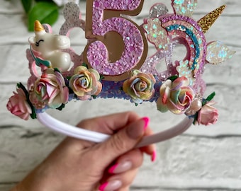 May include: A white headband with a pink glitter number 5, a rainbow, a unicorn, and pastel flowers. The headband is decorated with iridescent glitter.
