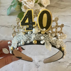 May include: A black and gold tiara with the number 40 on top. The crown is decorated with pearls, rhinestones, and gold accents. White leaves and a white ribbon are also included in the design.