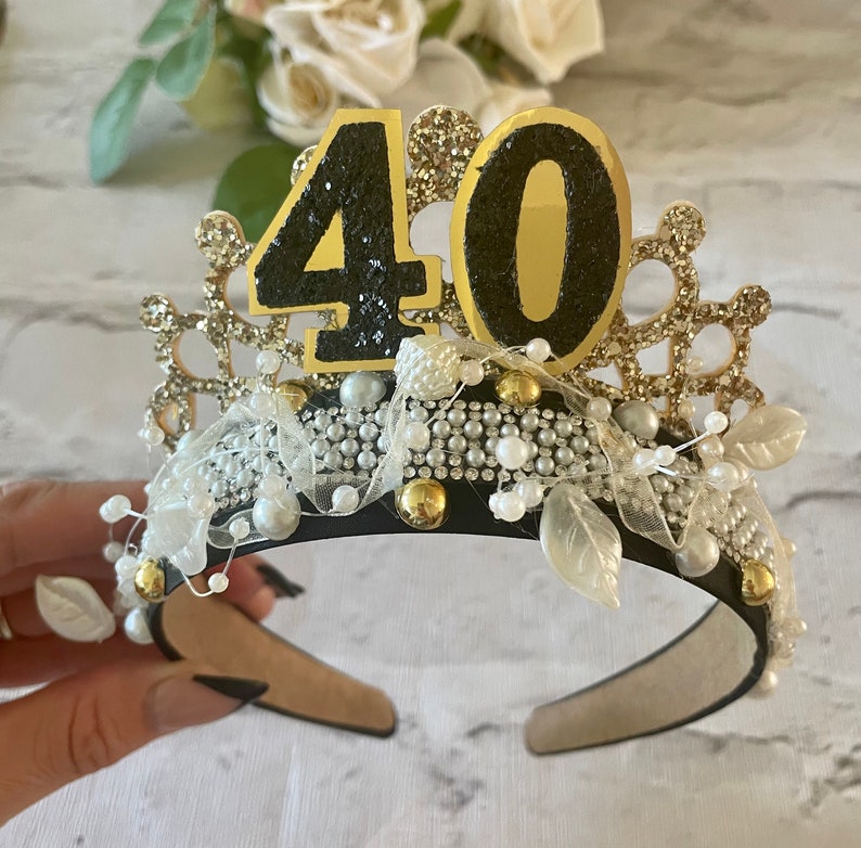May include: A black and gold tiara with the number 40 on top. The crown is decorated with pearls, rhinestones, and gold accents. White leaves and a white ribbon are also included in the design.