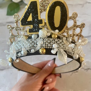 May include: A decorative crown with the number 40 on top. The crown is black with gold and silver accents, including pearls, glitter, and faux leaves. The crown is designed for a birthday celebration.