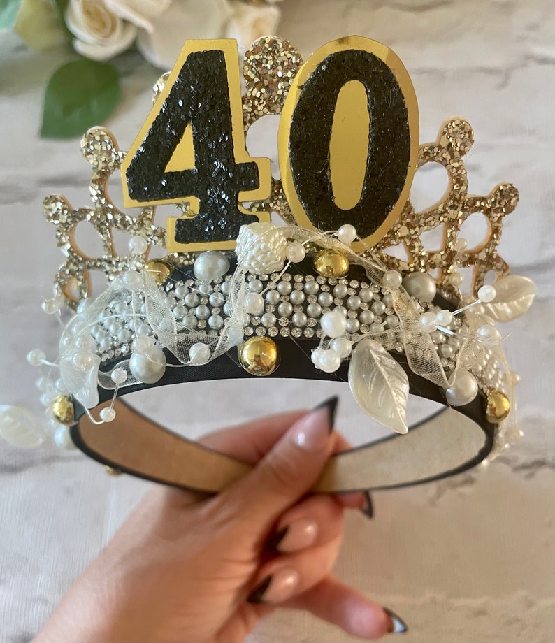 May include: A decorative crown with the number 40 on top. The crown is black with gold and silver accents, including pearls, glitter, and faux leaves. The crown is designed for a birthday celebration.