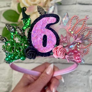 May include: A pink and green birthday crown with the number 6. The crown features glitter, rhinestones, and small flowers. The base is a pink headband.