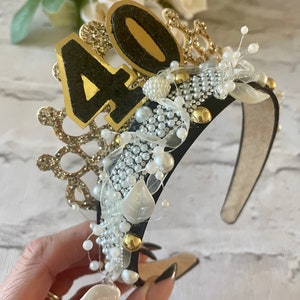 May include: A black headband with gold and white embellishments, including pearls, beads, and a crown design. The number "40" is prominently displayed in gold and black glitter. A festive accessory for a birthday celebration.