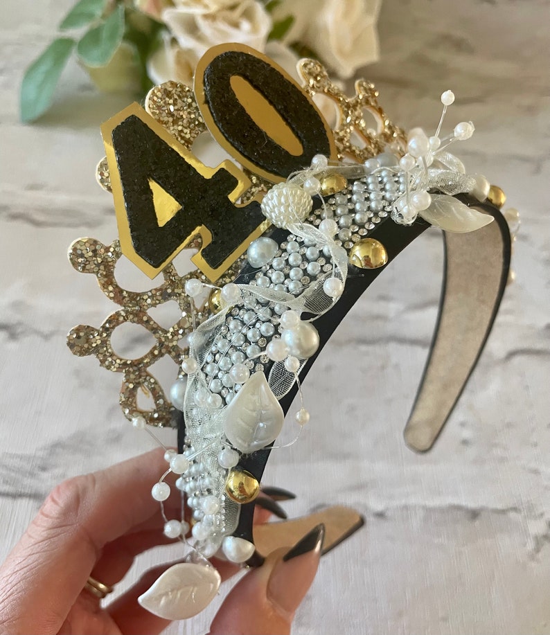 May include: A black headband with gold and white embellishments, including pearls, beads, and a crown design. The number "40" is prominently displayed in gold and black glitter. A festive accessory for a birthday celebration.