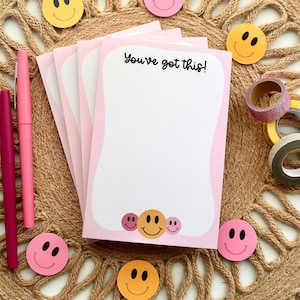 May include: A stack of pink and white notepads with a white background and pink border. The notepads have a smiley face design and the text "You've got this!" printed on the front.