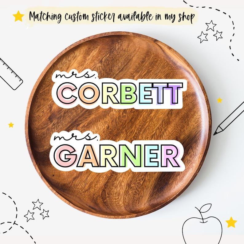May include: Two colorful teacher name stickers on a wooden plate. The top sticker says "mrs. CORBETT" in rainbow colors. The bottom sticker says "mrs. GARNER" in rainbow colors.