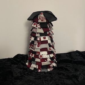 Puede incluir: Un adorno de árbol de Navidad de tela en capas decorado con tela granate, blanca y negra con el logotipo de Texas A&M. El árbol está coronado con un lazo negro y una joya granate.