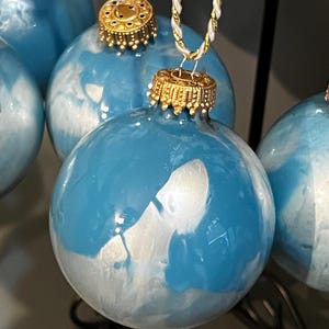 May include: Three blue and white Christmas baubles with gold accents. The baubles have a marbled effect with white swirls on a blue background. The baubles are hanging from gold cords.