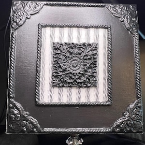 Puede incluir: Una caja decorativa cuadrada negra con detalles ornamentales en las esquinas y un borde similar a una cuerda. El centro presenta una tela a rayas y un diseño floral gris oscuro detallado. Un pomo transparente y facetado está adjunto en la parte inferior.