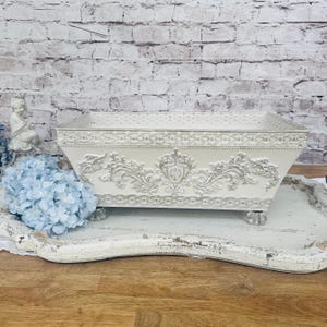 May include: A rectangular off-white metal planter box featuring intricate embossed scrollwork, a central crest design, and a decorative border along the top rim. The container is supported by four small, clear, faceted glass feet at the base.