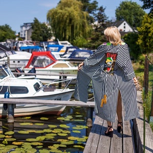 Könnte beinhalten: Eine Frau trägt eine schwarz-weiß gestreifte Robe mit einem bunten Patch auf dem Rücken, auf dem "DIVE" steht. Sie steht auf einem Holzsteg mit einem Yachthafen im Hintergrund.