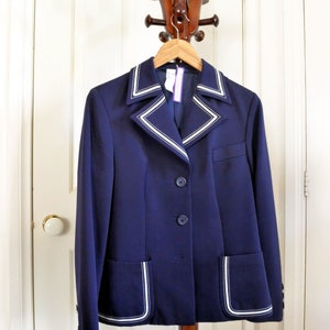 May include: A navy blue blazer with white piping around the collar, pockets, and cuffs. The blazer is hanging on a wooden coat rack.