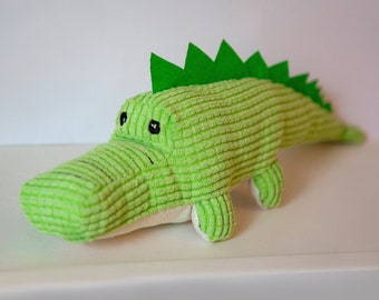 May include: A green plush alligator toy with a textured fabric and a green felt back fin. The alligator has black eyes and a white belly.
