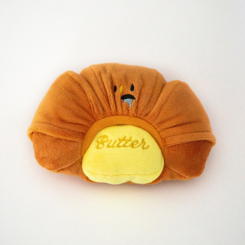 May include: A plush toy shaped like a croissant with a yellow face and the word "Butter" embroidered on the front. The toy is brown and yellow.