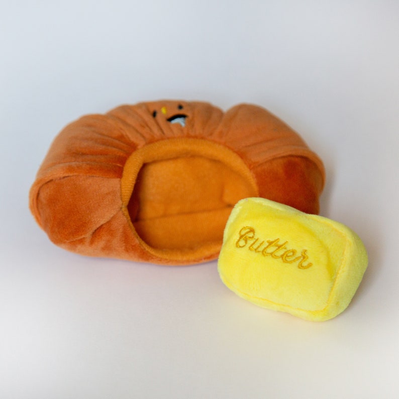 May include: A plush toy shaped like a stick of butter, with the word "Butter" embroidered on it. The butter is next to a plush toy shaped like a loaf of bread.