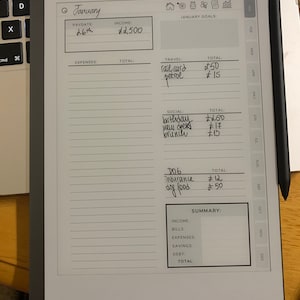 Puede incluir: Un planificador digital que muestra un presupuesto de enero con entradas manuscritas. El planificador muestra ingresos, gastos y objetivos, con un l&aacute;piz &oacute;ptico negro apoyado en el lateral. El planificador est&aacute; sobre una superficie de madera junto a un teclado de port&aacute;til.