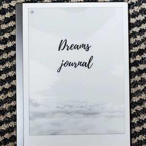 K&ouml;nnte beinhalten: Ein wei&szlig;er E-Reader mit schwarzem Rahmen und wei&szlig;em Hintergrund. Der Text "Dreams journal" ist in schwarzer Kursivschrift auf dem Bildschirm geschrieben. Der Bildschirm zeigt ein verschwommenes Bild eines bew&ouml;lkten Himmels.