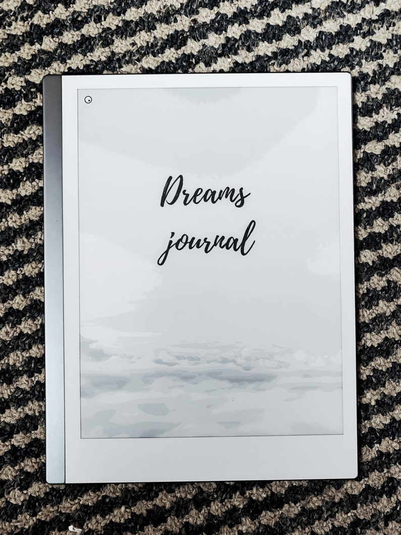 K&ouml;nnte beinhalten: Ein wei&szlig;er E-Reader mit schwarzem Rahmen und wei&szlig;em Hintergrund. Der Text "Dreams journal" ist in schwarzer Kursivschrift auf dem Bildschirm geschrieben. Der Bildschirm zeigt ein verschwommenes Bild eines bew&ouml;lkten Himmels.