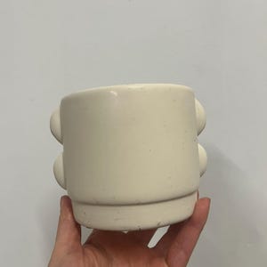 May include: Cream-coloured ceramic mug with two stacked, rounded handles. The mug has a cylindrical shape with a slightly flared base. The surface has a matte finish and appears to be handmade.