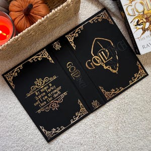 Könnte beinhalten: Schwarzer Buchumschlag mit goldfarbenen Buchstaben und dekorativen Eckdesigns. Der Umschlag trägt die Aufschrift "GOLDINCH" und enthält das Zitat "My name is Auren Turley and I am stronger than the dark."