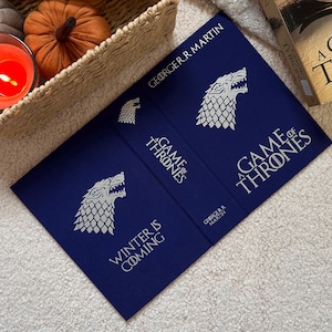 May include: A Game of Thrones book cover in navy blue with white text and a direwolf emblem. The cover features the author's name, the book title, and the phrase "Winter is Coming."
