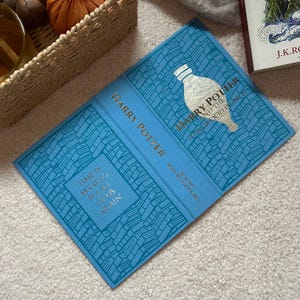 Könnte beinhalten: Ein blaues Hardcover-Buch mit dem Titel "Harry Potter und der Halbblutprinz" mit goldener Schrift und einer silbernen Illustration. Auf dem Cover steht das Zitat "Die Zeit macht uns wieder zu Narren."