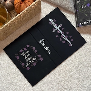 May include: Black book cover featuring the word "Powerless" in white script. A silver dagger with purple floral accents is on the right. The left side has a floral frame with the text "Vicious Little Thing Isn't She".