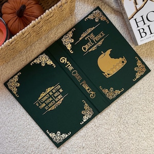 Può includere: Libro aperto intitolato "The Cruel Prince" con copertina verde scuro e scritte e dettagli dorati. La copertina presenta un design a mela e corona dorata. Il libro è aperto su una pagina con una citazione. Un cesto con zucche è sullo sfondo.