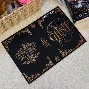 Könnte beinhalten: Schwarzer Buchumschlag mit goldfarbenem Folienschriftzug und dekorativen Eckdesigns. Das Wort "GLINT" ist prominent dargestellt, zusammen mit dem Zitat "Manchmal müssen Dinge zuerst ruiniert werden, um dann neu gemacht zu werden."