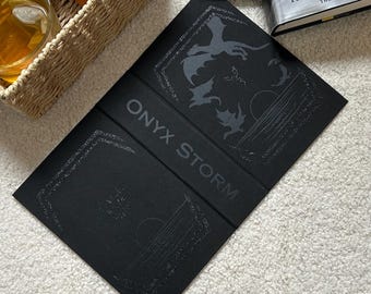 Puede incluir: Cubierta de libro negra con el título "ONYX STORM" en letras grises. La cubierta presenta ilustraciones de dragones y una escena celestial. El libro está abierto, revelando las páginas interiores. Un libro de tapa dura es visible en el fondo.
