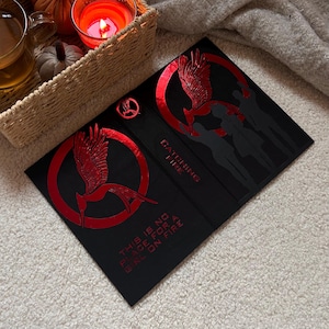 May include: Black book with red foil accents featuring a stylized bird within a circle, and the text "Catching Fire". The book also has the text "This is no place for a girl on fire".