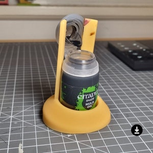 Op de afbeelding: Een gele plastic flessenhouder met een doorzichtige plastic fles erin. De fles is gelabeld met "Citadel Shade Nuln Oil".