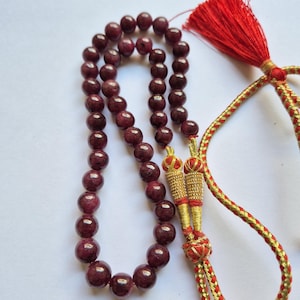 Puede incluir: Un collar de cuentas rojo oscuro, probablemente piedras preciosas. Una borla roja y un cordón dorado y rojo completan el diseño. Las cuentas son redondas y pulidas, reflejando la luz. El collar se utiliza probablemente para la oración o la meditación.