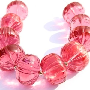 May include: A pink beaded necklace with large, round, transparent beads. Each bead has a ribbed texture and is strung together with gold-colored connectors. The necklace is displayed on a white surface, with the beads reflecting light.