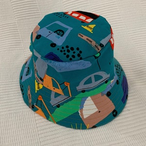 May include: A teal blue bucket hat with a colorful print of cars, trucks, and construction equipment. The hat has a wide brim and a flat crown.