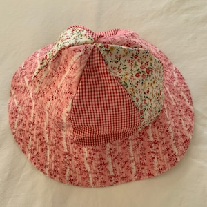 May include: A pink and red patchwork sun hat with a floral print. The hat is made of cotton fabric and has a wide brim.