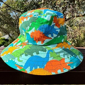 May include: A blue and green bucket hat with a dinosaur print. The hat has a wide brim and is made of a lightweight fabric.