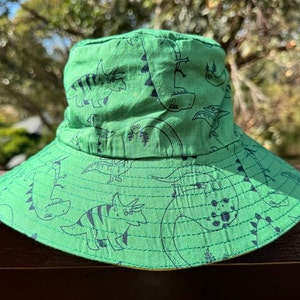 May include: Green bucket hat with a dinosaur print. The hat has a wide brim and a flat crown.