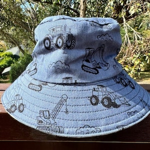 May include: A blue denim bucket hat with a black and white construction vehicle print. The hat features tractors, excavators, and cranes.